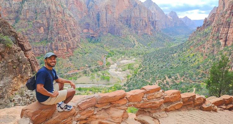 Randonneur souriant agenouillé sur un rebord de roche rouge surplombant une vallée de canyon spectaculaire.