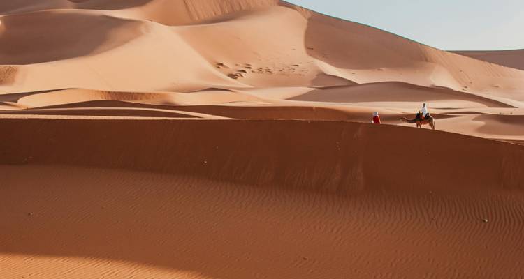 Weite gewellte Dünenlandschaft mit zwei Reitern, einer auf einem Kamel, winzig vor endlosem Sand.