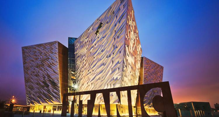 La fachada angular e iluminada del museo Titanic Belfast resplandeciendo contra un cielo crepuscular de color azul profundo.