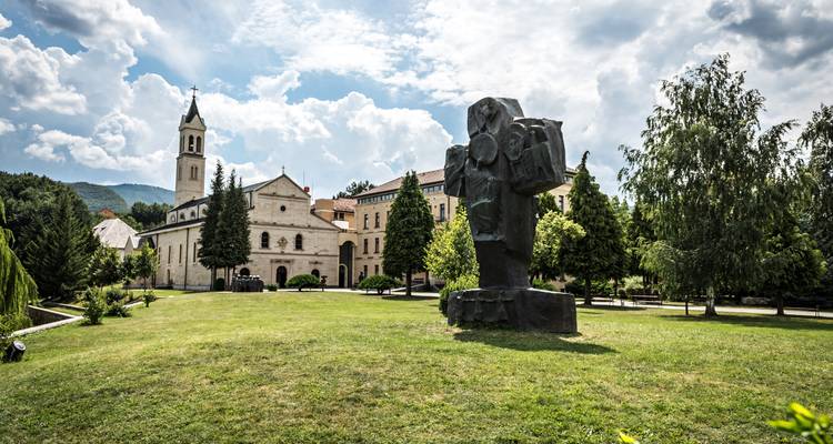Klosteranlage mit Glockenturm und moderner Skulptur inmitten gepflegter Gärten unter dramatischem Himmel