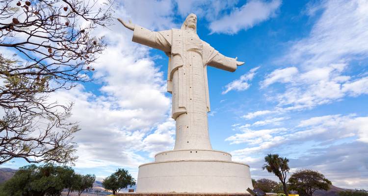 Torenhoge witte standbeeld van Christus met uitgestrekte armen tegen een heldere blauwe lucht en verspreide wolken