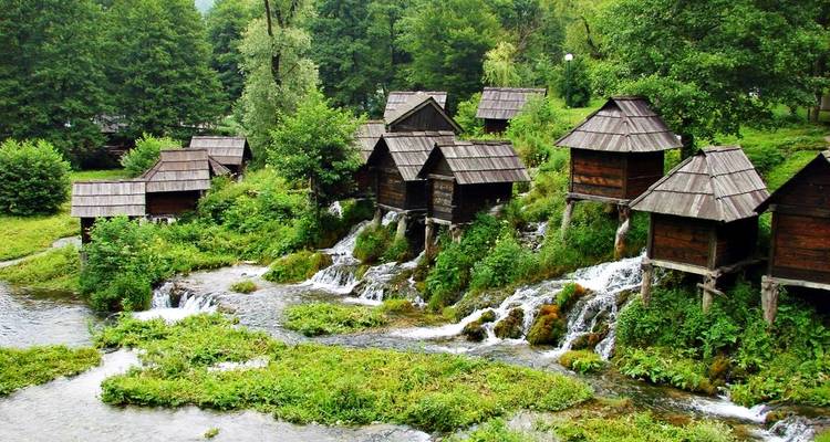 Fila de molinos de agua tradicionales de madera encaramados sobre pequeñas cascadas en un entorno ribereño verdoso.
