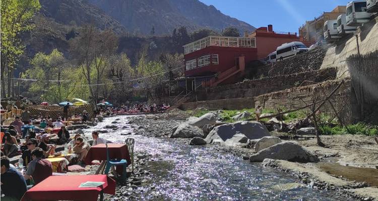 Crowded riverside cafés with colourful umbrellas line a rocky stream at the base of mountains.