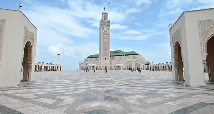 De torenhoge minaret van de Hassan II-moskee domineert een uitgestrekt gepatroneerd plein onder een heldere hemel in Casablanca.