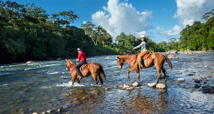 Jinetes a caballo cruzan río rocoso poco profundo en medio de bosque tropical bajo cielo azul brillante