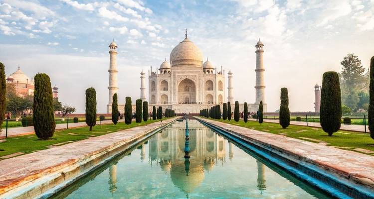 Ikonischer Blick auf das Taj Mahal, das sich im langen, von Zypressen gesäumten Wasserbecken unter einem blau-bewölkten Himmel spiegelt