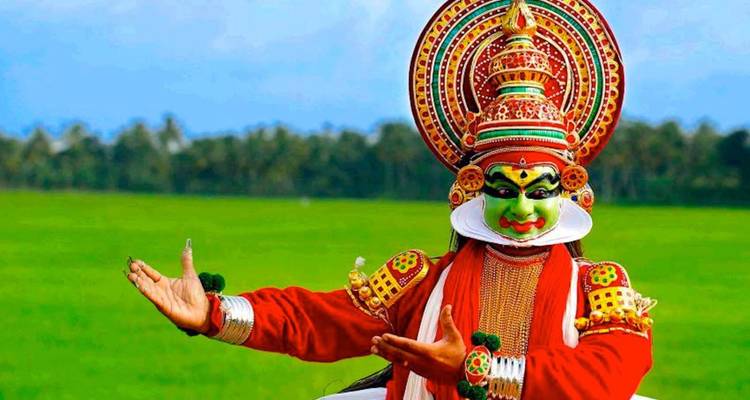Kathakali-uitvoerder in uitbundig kostuum en make-up gebarend tegen groene rijstvelden.