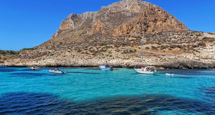 Aguas turquesas salpicadas de pequeñas embarcaciones junto a una isla rocosa escarpada bajo un cielo azul despejado.