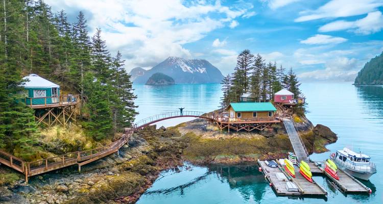 Un îlot forestier isolé avec des cabanes en bois et une passerelle courbe se dresse dans les eaux turquoise du fjord avec des pics enneigés au-delà.