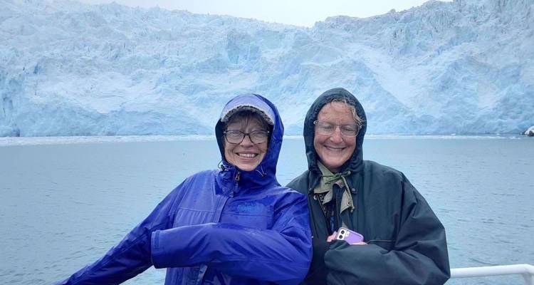 Deux femmes souriantes en vestes de pluie posent sur un bateau avec un glacier bleu massif derrière elles.