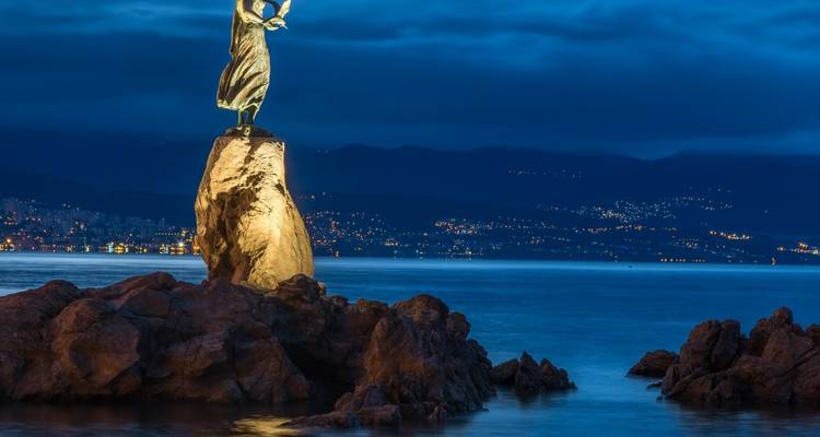 La Doncella con la Gaviota, estatua iluminada sobre una roca encima del Adriático en calma durante la hora azul, Opatija a la distancia.