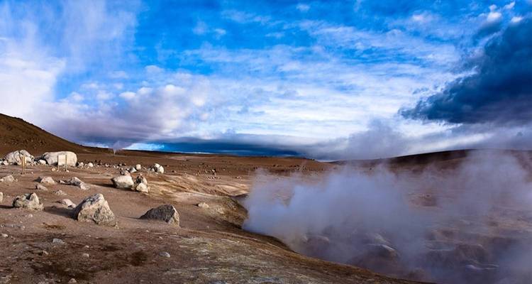 De la vapeur s'élève d'un champ de geysers en haute altitude sous un ciel bleu dramatique et des nuages ondulants.