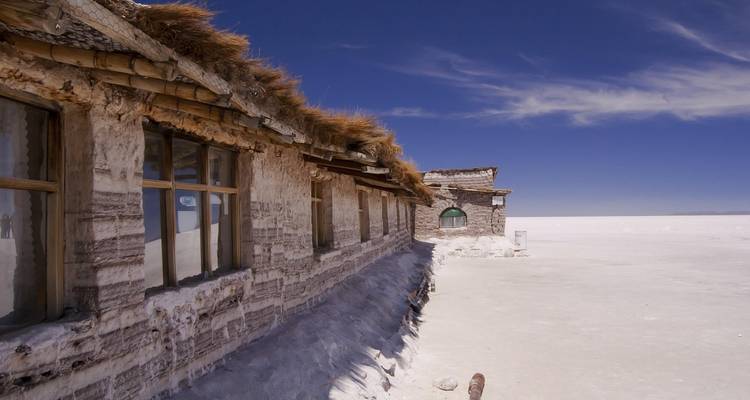 Un lodge rustique construit de blocs de sel se dresse seul sur la vaste plaine blanche sous un ciel bleu profond.