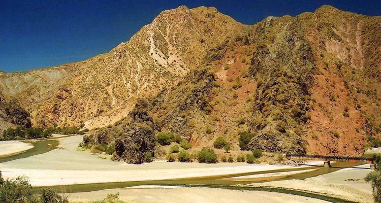 Une rivière sinueuse traverse des montagnes arides multicolores et des berges sableuses sous un ciel dégagé.