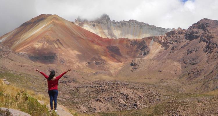 Une voyageuse lève les bras en signe de triomphe devant les montagnes andines colorées striées de minéraux.