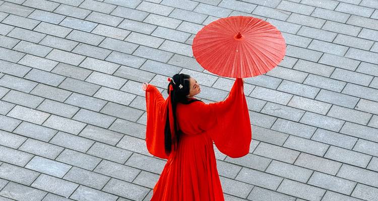 Woman in flowing red traditional dress holding a matching parasol while walking across grey stone tiles
