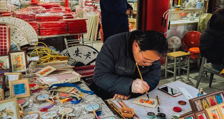 An artisan paints traditional designs at a bustling Chinese market stall full of souvenirs and fans.