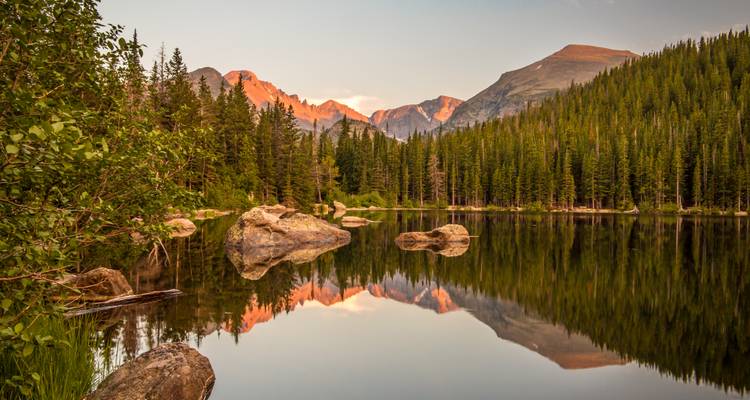 Lac de montagne calme reflétant la forêt de pins et les pics aux reflets rosés au coucher du soleil