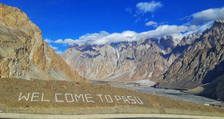 "WELCOME TO PASU" written in white stones before the dramatic Passu Cones mountains