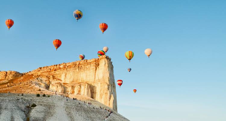 Des montgolfières colorées s'élèvent dans un ciel bleu dégagé à côté d'une falaise blanche imposante en Cappadoce.
