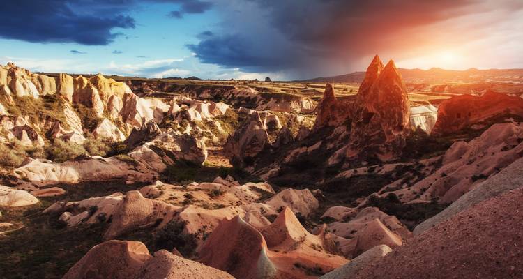 La lumière du coucher de soleil baigne les vallées rugueuses aux couleurs rosées et les flèches rocheuses déchiquetées de la Cappadoce sous un ciel dramatique.