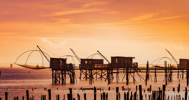 Silhouettierte hölzerne Fischerhütten auf Stelzen mit traditionellen Netzen entlang der Küste bei glühendem orangefarbenem Sonnenuntergang.