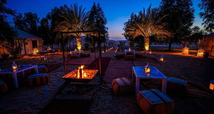 Campement atmosphérique du désert au crépuscule avec palmiers, lanternes et un foyer lumineux installé sur des tapis à motifs.