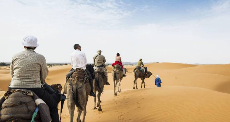 Voyageurs montés sur des chameaux en file indienne traversant des dunes dorées du désert sous un ciel bleu pâle.