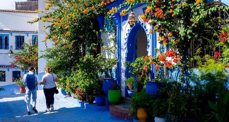 Deux touristes marchent le long d'une ruelle pittoresque peinte en bleu, ornée de fleurs éclatantes et de pots.