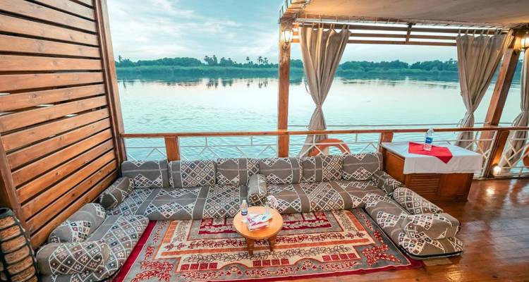 Coin salon confortable avec coussins sur le pont en bois d'un bateau fluvial avec vue sur le Nil au coucher du soleil.