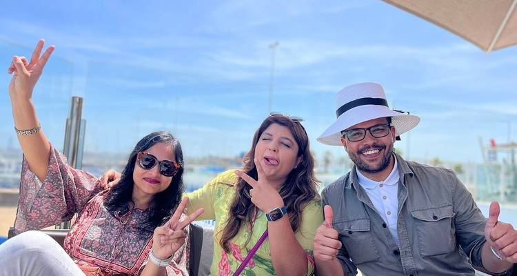 Drie vrienden poseren met vredestekens en duimen omhoog bij een buitencafé onder een blauwe hemel