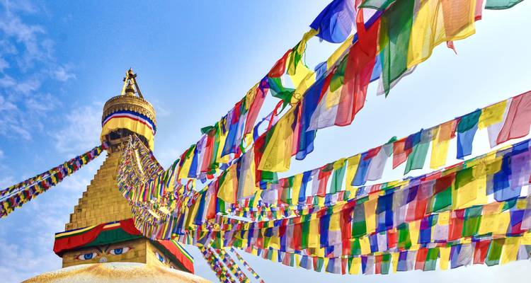 Bunte Gebetsfahnen wehen von der goldenen Stupa, die mit Buddha-Augen gekrönt ist, vor einem strahlend hellen Himmel.