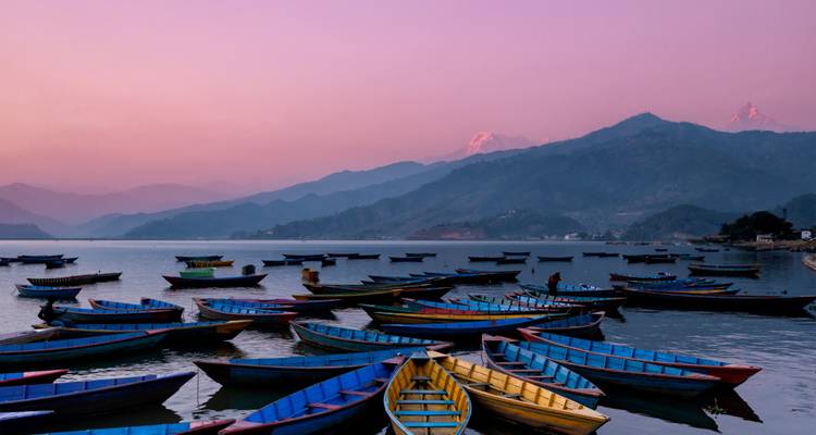 Lebhaft bemalte Holzboote, die bei Sonnenaufgang auf einem ruhigen See vor dem Hintergrund dunstiger Himalaya-Gipfel vor Anker liegen.