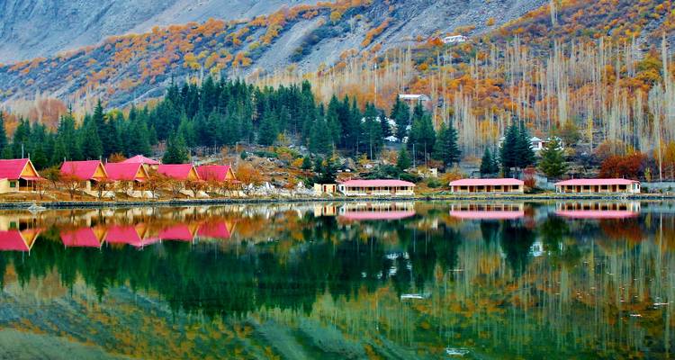 Fila de cabañas de techo rojo junto al lago y bosque otoñal reflejados en agua turquesa en Skardu