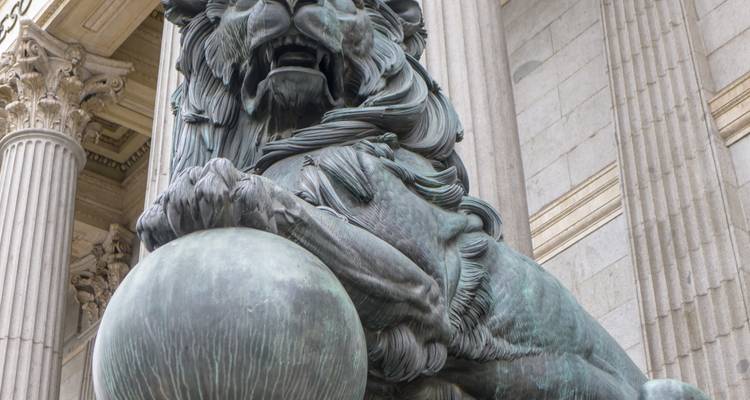 Close-up of bronze lion statue guarding entrance of a neoclassical building with marble columns.