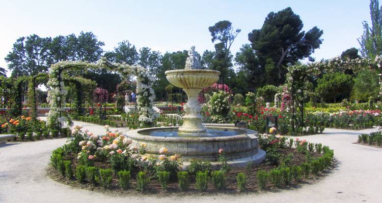 Stone fountain surrounded by manicured rose beds and arched trellises in city park.