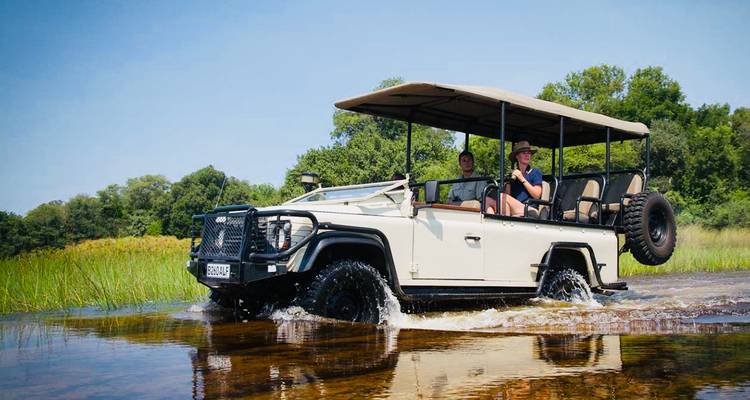 Safariauto spettert door ondiep water omringd door weelderige groene graslanden en bomen.