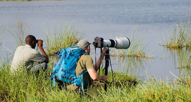 Twee fotografen hurken bij de rand van een rustig meer en gebruiken een verrekijker en een grote telelens.