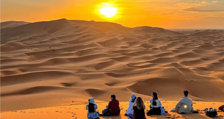 Reizigers zitten bovenop een duin en kijken naar de zon die ondergaat boven de golvende woestijnzanden.