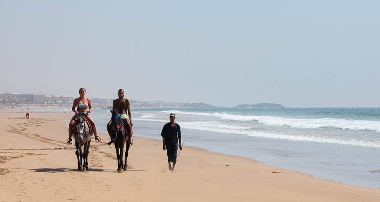 Two riders on horseback and a local guide walking along a vast sandy Atlantic beach