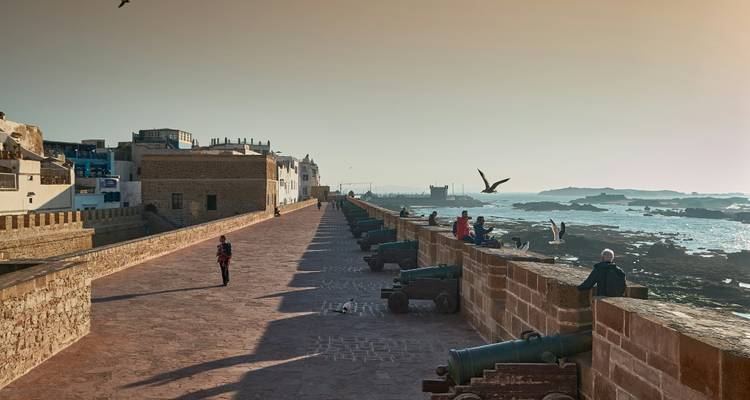 Stone ramparts with old cannons facing the Atlantic at sunset in Essaouira while people stroll and seagulls soar