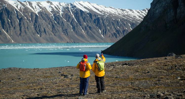 Dos excursionistas señalan hacia el otro lado de un fiordo glacial de aguas turquesas en dirección a crestas nevadas y empinadas.