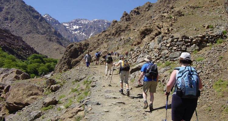 Wandelaars die een rotsachtig bergpad volgen onder een heldere hemel met besneeuwde toppen in de verte.