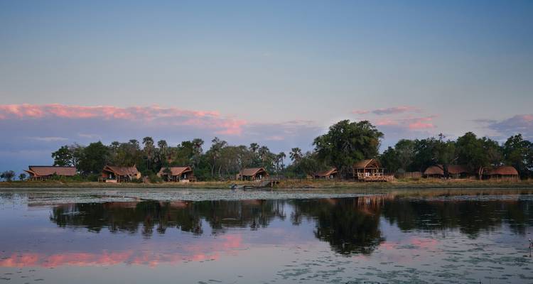 Chalets de lodge nichés parmi les arbres se reflétant dans une rivière sereine au crépuscule avec des nuages roses au-dessus.