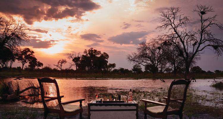 Deux chaises et une table basse disposées pour l'apéritif au bord d'une rivière calme sous un ciel de coucher de soleil dramatique.