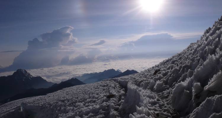 Ijsbedekte bergkam boven een zee van wolken met felle zon en ijsformaties op de voorgrond.