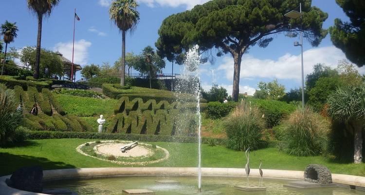 Manicured public garden with fountain, sculpted hedges and tall umbrella pine against a blue sky.