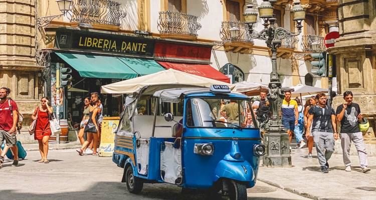 Colorful street scene in an old Italian city with a bright blue three-wheeler tuk-tuk parked in front of the bookshop “Libreria Dante” while pedestrians walk by historic stone buildings.