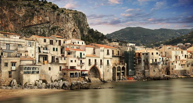 Picturesque coastal village with pastel stone houses clinging to a rocky shoreline under a dramatic sky, backed by rugged hills and calm turquoise sea.