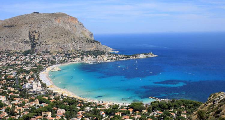 A sweeping aerial view over a turquoise bay lined with sandy beaches and a hillside city, framed by a rugged cliff and deep blue Mediterranean Sea.